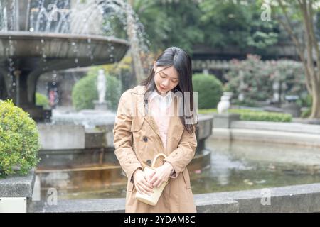 Eine langhaarige taiwanesische Frau im Alter von 20 Jahren in einem beigefarbenen Trenchcoat verbringt ihren Winternachmittag an einem Brunnen in einem Park im Bezirk Xinyi, Taipeh Stockfoto