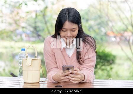 Eine langhaarige taiwanesische Frau im Alter von 20 Jahren, die einen rosafarbenen Pullover trägt, bedient ein Smartphone, während sie auf einer Parkbank im Bezirk Xinyi, Taipei, Ta, sitzt Stockfoto