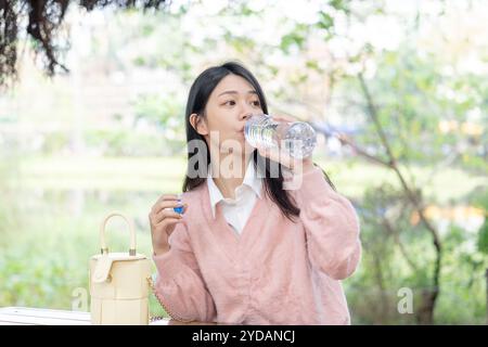 Eine langhaarige taiwanesische Frau im Alter von 20 Jahren mit rosa Pullover sitzt auf einer Parkbank im Bezirk Xinyi, Taipeh, Taiwan und trinkt Wasser aus einem Plastik Stockfoto