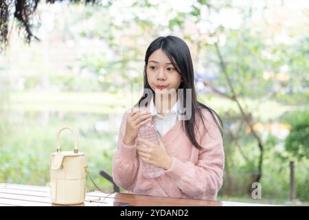 Eine langhaarige taiwanesische Frau im Alter von 20 Jahren mit rosa Pullover sitzt auf einer Parkbank im Bezirk Xinyi, Taipeh, Taiwan und trinkt Wasser aus einem Plastik Stockfoto