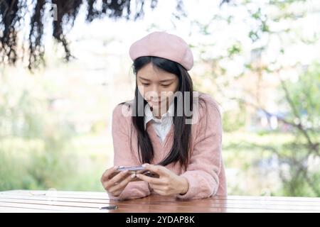 Eine langhaarige taiwanesische Frau im Alter von 20 Jahren, die einen rosafarbenen Pullover trägt, bedient ein Smartphone, während sie auf einer Parkbank im Bezirk Xinyi, Taipei, Ta, sitzt Stockfoto