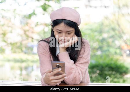 Eine langhaarige taiwanesische Frau im Alter von 20 Jahren, die einen rosafarbenen Pullover trägt, bedient ein Smartphone, während sie auf einer Parkbank im Bezirk Xinyi, Taipei, Ta, sitzt Stockfoto