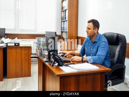 Junger Mann in Denim-Hemd sitzt am Schreibtisch und arbeitet am PC im Büro. Der Mitarbeiter macht seine Arbeit am Arbeitsplatz. Stockfoto