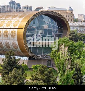 Seitenansicht des Teppichmuseums in Baku, Aserbaidschan, zeigt seine beeindruckende moderne Architektur mit einem geschwungenen goldenen großen Fenster, eingebettet in üppiges Grün Stockfoto