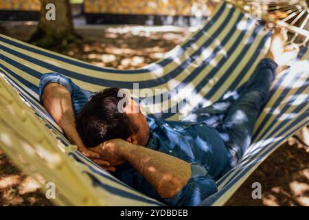 Ein junger Mann in lässiger Kleidung liegt in einer Hängematte im Hinterhof des Hauses. Ein Mann entspannt sich auf der Terrasse im Schatten der Bäume. Eine Person Stockfoto