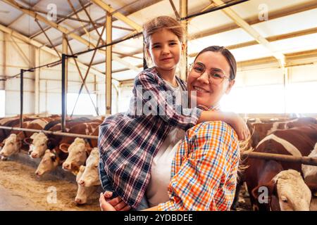 Eine junge Frau in lässiger Kleidung hält ihre kleine Tochter in den Armen in einem Kuhstall. Eine glückliche Bauernfamilie in einer Scheune. Stockfoto