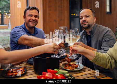 Junge Leute lachen und stoßen draußen auf der Terrasse an. Fröhliche Freunde feiern das Treffen und trinken Champagner. Männer zünden die Gläser und essen einen Stockfoto