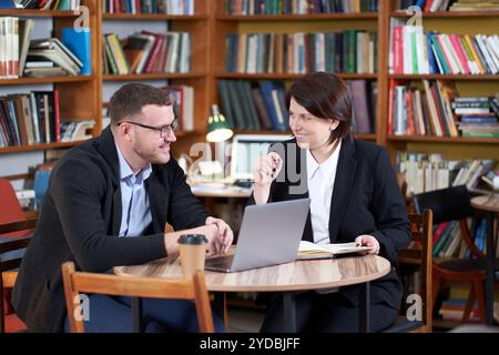 Mann und Frau, die mit einem Laptop arbeiten, sitzen in einem modernen, stilvollen Zimmer mit Bücherregalen im Hintergrund in Bibliothek oder Buchladen Stockfoto