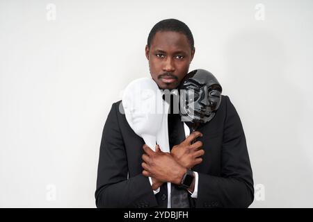 Afrikanischer junger Mann mit schwarzem Anzug, der weiße und schwarze Plastikmasken hält und das Gesicht enthüllt Stockfoto