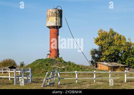 Alter Wasserturm auf dem Land. Ein rostiger, veralteter Wasserturm steht auf einem offenen Feld mit einem beschädigten Zaun und einem kleinen Ziegelschuppen daneben. Die Stockfoto