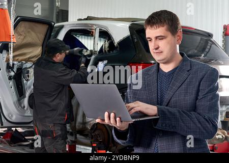 Manager Laptop halten, während in der Nähe Auto in der Werkstatt mit Mechaniker Reparatur Auto auf Hintergrund stehen Stockfoto