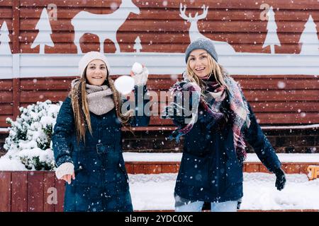 Zwei junge, fröhlich lächelnde kaukasische Frauen im Schneefall werfen Schneebälle auf die Kamera. Stockfoto