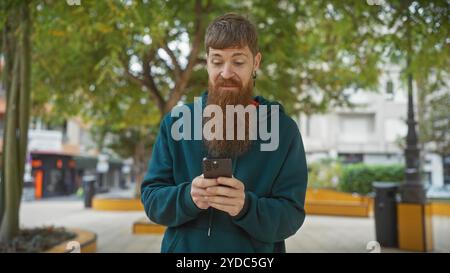 Hübscher rothaariger Mann mit Bart mit Smartphone im Park, in Bäumen und im Hintergrund der Stadt. Stockfoto