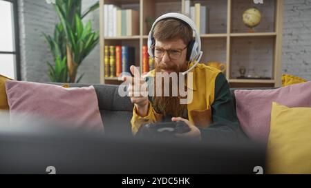 Bärtiger junger Mann in Brillen und Kopfhörern, der drinnen spielt und mit Begeisterung zeigt Stockfoto