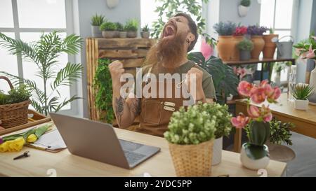 Aufgeregter bärtiger Mann in Florismusschürze jubelt zwischen Laptop und lebendigen Pflanzen drinnen. Stockfoto