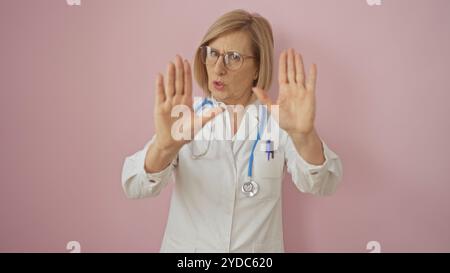 Reife Ärztin mit blonden Haaren und Brille, die aufhört, mit weißem Mantel und Stethoskop, isoliert vor rosa Hintergrund. Stockfoto
