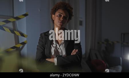 Selbstbewusste Frau mit überkreuzten Armen an einem drinnen Tatort, mit ernstem Ausdruck und Detektivambiente. Stockfoto