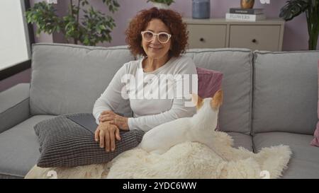 Eine ältere kaukasische Frau mit lockigem Haar und Brille sitzt bequem auf einem Sofa mit ihrem Haustier chihuahua in einem gemütlichen, gut dekorierten Wohnzimmer. Stockfoto