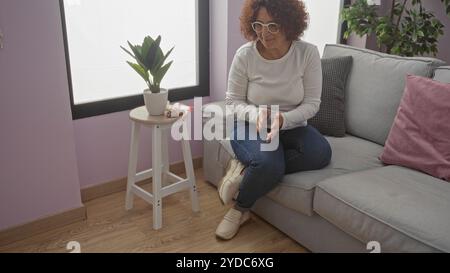 Eine ältere kaukasische Frau mit lockigen Haaren sitzt in einem gemütlichen Wohnzimmer mit Pflanzen und minimalistischem Dekor um sie herum. Stockfoto