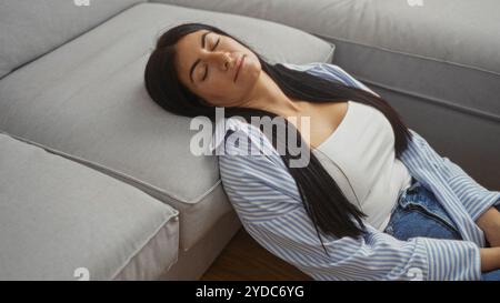 Eine junge hispanische Frau entspannt sich und sitzt im Wohnzimmer ihres Hauses, trägt legere Kleidung mit geschlossenen Augen und lehnt sich an ein Grau Stockfoto