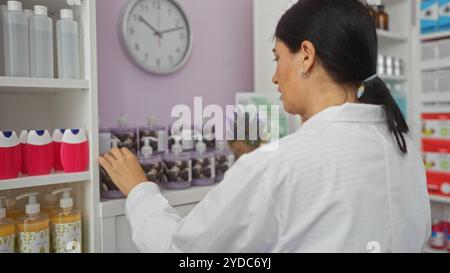 Eine junge hispanische Frau mit brünetten Haaren arrangiert Produkte in einer Apotheke, umgeben von verschiedenen Flaschen und Spendern, mit einer Uhr an der Wand Indi Stockfoto