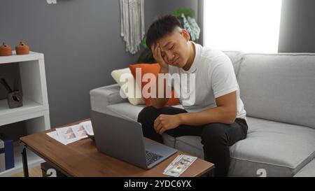Junger Mann, der im Wohnzimmer sitzt, mit Hand auf der Stirn, der über Laptop gestresst schaut, mit uns Dollars und Dokumenten auf dem Tisch Stockfoto