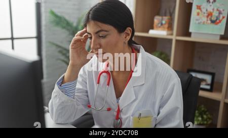 Eine junge brünette hispanische Frau in einem Krankenhauszimmer, die einen Arztmantel und ein Stethoskop trägt, sieht konzentriert aus, während sie an einem Computer arbeitet und hebt hervor Stockfoto