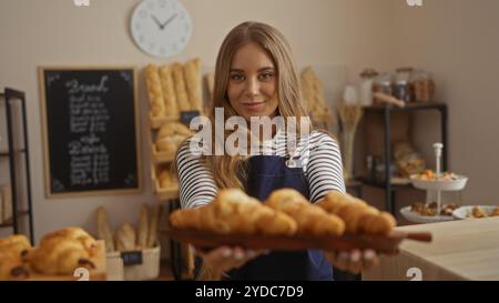Junge blonde Frau mit Schürze in der Bäckerei mit Tablett mit frisch gebackenen Croissants, mit Regalen mit Brot und Gebäck im Hintergrund sichtbar Stockfoto