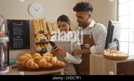Mann und Frau, die Schürzen in einer Bäckerei tragen und gemeinsam ein Telefon überprüfen, umgeben von frischem Brot und Gebäck Stockfoto