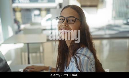 Junge Frau lächelt in einem Büro mit einem Laptop und zeigt eine helle Arbeitsumgebung in Innenräumen mit natürlichem Licht durch die Fenster, die auf ein P Stockfoto