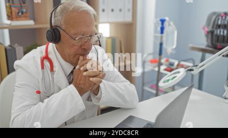 Ein älterer kaukasier in einer Klinik, der Kopfhörer und einen weißen Mantel trägt, sitzt vor einem Laptop und erscheint tief in Gedanken in einem professionellen Arzt Stockfoto