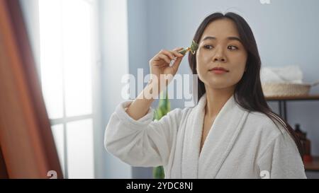 Asiatische Frau im Spa, die Jadewalze zur Hautpflege verwendet, während sie einen weißen Bademantel in einem Wellnessraum mit weichem Tageslicht trägt. Stockfoto