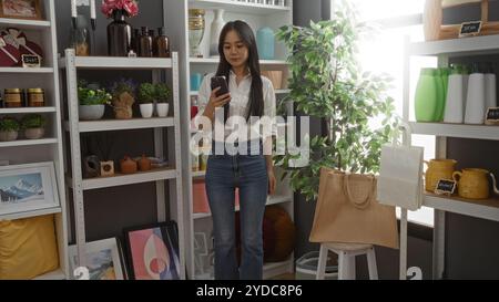 Junge chinesin, die auf ihrem Handy surft, während sie in einem wunderschön dekorierten Haus-Dekorationsgeschäft steht, gefüllt mit verschiedenen Ziergegenständen. Stockfoto