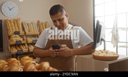 Junger hispanischer Mann in brauner Schürze mit Smartphone im Bäckereiinnenraum mit Brotregalen im Hintergrund Stockfoto