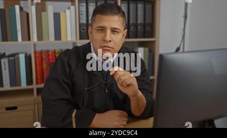 Junger hispanischer Mann in Richterbekleidung, der in einem Büro sitzt, mit Regalen voller Bücher und Ordner Stockfoto