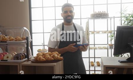 Hübscher junger Mann, der drinnen in einer Bäckerei mit Gebäck arbeitet, während er eine Tablette hält und eine gestreifte Schürze trägt. Stockfoto
