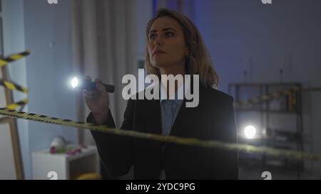 Eine ernsthafte junge blonde Frau untersucht einen Tatort drinnen, beleuchtet von ihrer Taschenlampe. Stockfoto