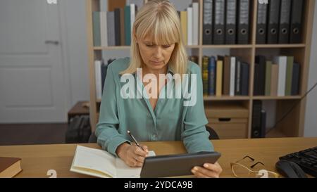 Frau mittleren Alters, die in einem Büro auf einem Tablet arbeitet, während sie Notizen in einem Buch macht Stockfoto