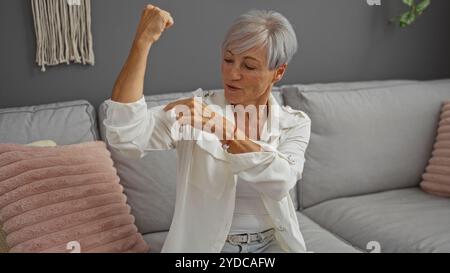 Ältere Frau, die ihren Arm in einem Wohnzimmer beugte, Stärke und Selbstvertrauen zeigte, auf einem Sofa mit grauem und rosa Dekor, kurzen grauen Haaren und weißem weiß saß Stockfoto