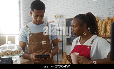 Mann, der ein Tablet benutzt, und Frau, die Kaffee in einer Bäckerei hält, beide Schürzen tragen, besprechen geschäftliche Aufgaben in einem Innengeschäft. Stockfoto