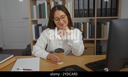 Eine junge Frau in einem Büro, die eine Kaffeetasse hält, an einem Schreibtisch mit Büchern und einem Computer, umgeben von Bücherregalen Stockfoto