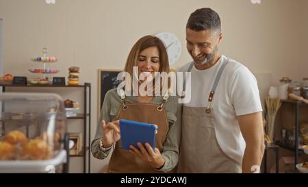 Mann und Frau tragen Schürzen, die zusammen eine Tablette in einer gemütlichen Bäckerei verwenden, mit Donuts und Gebäck im Hintergrund, die als glückliches Busin erscheinen Stockfoto