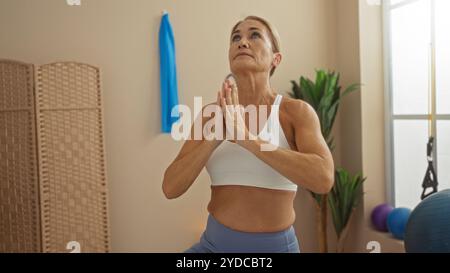 Reife Frau, die Yoga in einem Indoor-Sportzentrum praktiziert, trägt einen weißen Sport-BH und blaue Leggings, fokussiert mit Händen in Gebetsposition. Stockfoto