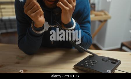 Ein Mann mit Handschellen, der ein Telefon in einer Polizeiwache benutzt und eine angespannte rechtliche Situation darstellt. Stockfoto