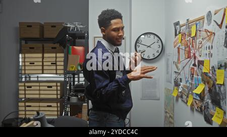 Ein afroamerikanischer Mann analysiert Beweise in einem Detektivbüro, das mit Fotos und einem Whiteboard überfüllt ist. Stockfoto