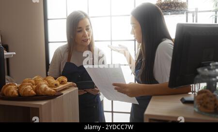 Bäckerinnen diskutieren ihre Arbeit in einer Bäckerei im Innenbereich, umgeben von frischem Gebäck, und betonen Teamarbeit und Zusammenarbeit in einer Ladenumgebung. Stockfoto