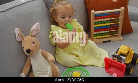 Das niedliche Kleinkind in einem gelben Kleid sitzt zu Hause auf einer Wohnzimmercouch, umgeben von Spielzeug wie einem Plüschkänguru, Abacus und einem Spielzeugwagen Stockfoto