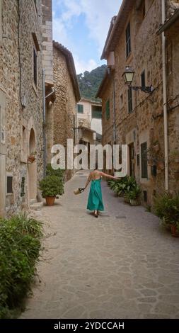 Frau, die durch die charmante Kopfsteinpflasterstraße in valldemossa, mallorca, läuft, in türkisfarbenem Kleid, umgeben von rustikalen Steinhäusern und mediter Stockfoto