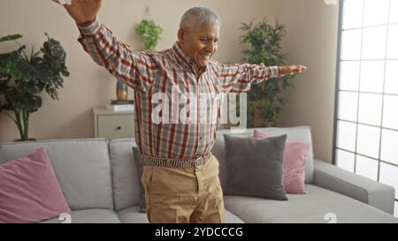 Ein hispanischer Mann mittleren Alters mit grauen Haaren, gekleidet in einem karierten Hemd, steht mit ausgestreckten Armen in einem gemütlichen Wohnzimmer neben einer grauen Couch und Hous Stockfoto