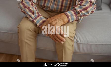 Ein älterer Mann, der in einem Wohnzimmer sitzt und sein Handgelenk vor Schmerzen hält und sein kariertes Hemd und seine beige Hose hervorhebt. Stockfoto